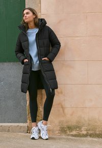 Abrigo acolchado negro con capucha, cierre frontal y dos bolsillos laterales. Lleva puesto un sudadera azul claro y leggings negros, combinados con zapatillas blancas.