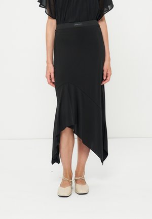 Kvinde iført en sort asymmetrisk midi-nederdel med flæsekant, kombineret med beige snøreløse flade sko, stående mod en hvid baggrund.