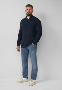 Marine blauer Reißverschluss-Pullover mit strukturiertem Stoff, Stehkragen und einem kleinen Logo-Patch, kombiniert mit hellblauen Jeans und weißen Sneakers.
