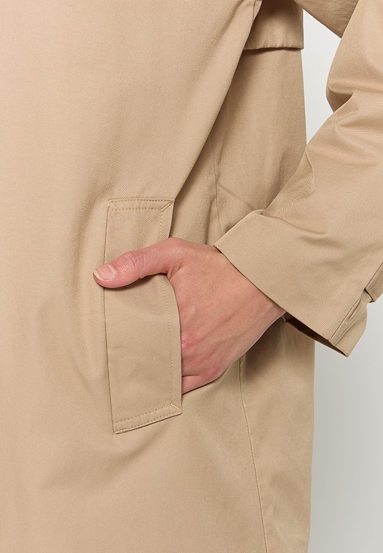 Trench coat beige con una texture liscia, dotato di tasca laterale e maniche lunghe. Il tessuto sembra leggero e resistente all'acqua.