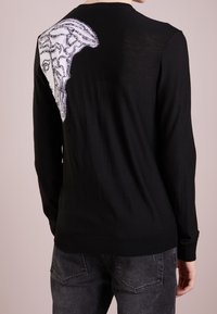 Svart stickad tröja med en stor vit grafisk design på vänster axel. Långa ärmar och ribbad mudd ger textur.