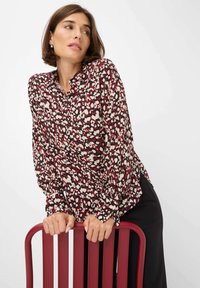 Langärmlige Bluse mit einem maronenfarbenen, cremefarbenen und schwarzen abstrakten Muster, ausgestattet mit einem Hemdkragen und gerafften Ärmeln. Hoch taillierte schwarze Hose.