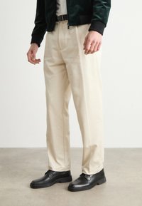 Pantalones de pierna ancha color crema con una textura suave, combinados con zapatos de cuero negros. El conjunto incluye una chaqueta bomber verde oscuro.