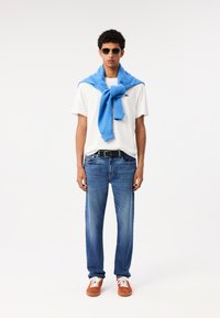T-shirt bianca, maglione blu annodato intorno al collo, jeans blu e sneakers arancioni. Modello che indossa occhiali da sole, in piedi davanti a uno sfondo bianco.