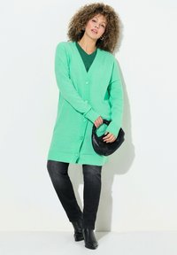 Ljusgrön lång kofta med knappar, bärs över en mörkgrön mönstrad topp, ihop med svarta jeans och en svart handväska.