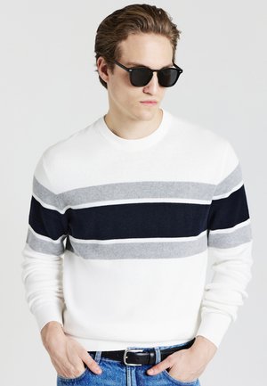 Maglione bianco con strisce navy, grigie e grigio chiaro; scollatura rotonda, polsini e orlo a costine; indossato con occhiali da sole scuri e jeans di denim.