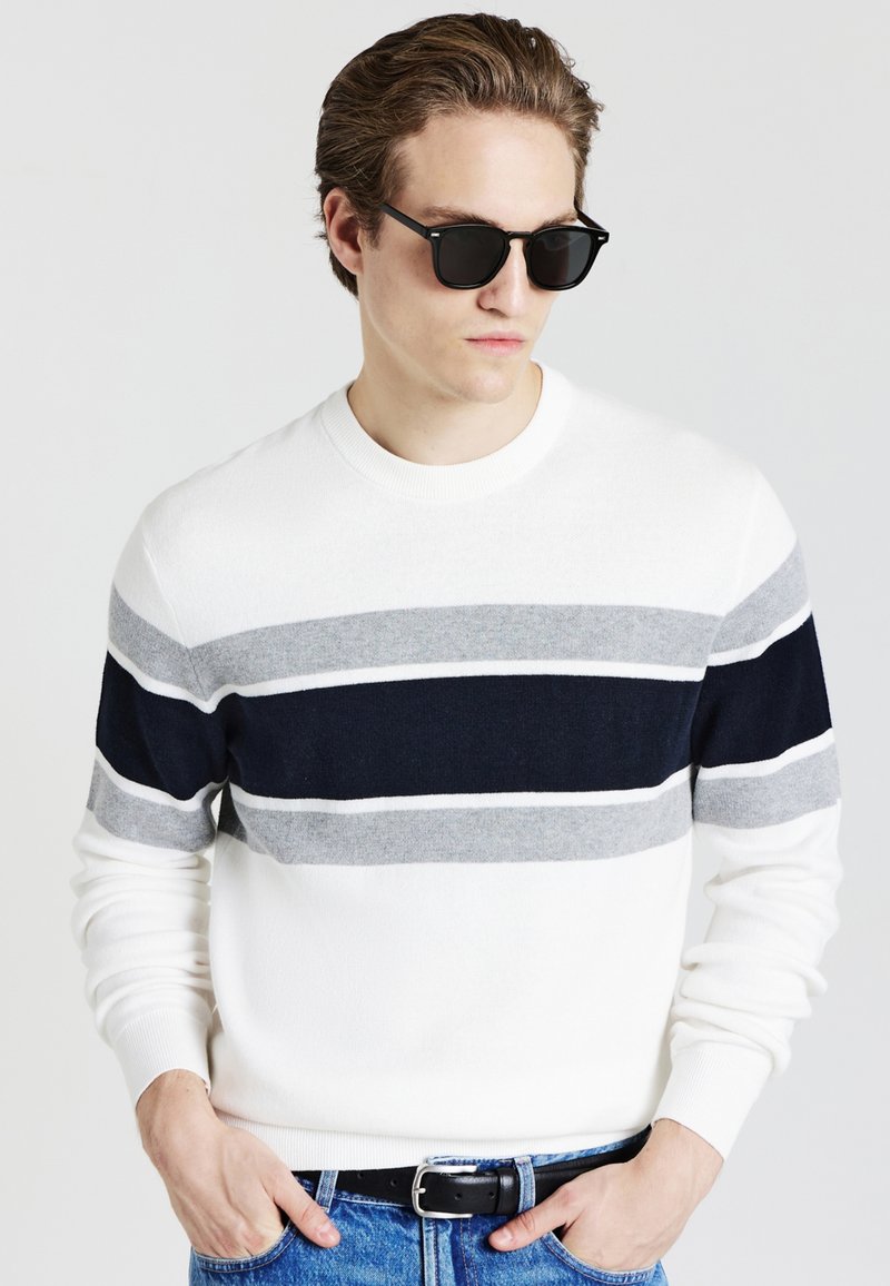 Weißer Pullover mit Streifen in Marineblau, Grau und Hellgrau; runder Ausschnitt, gerippte Bündchen und Saum; getragen mit dunklen Sonnenbrillen und Jeans.