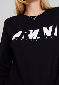 Svart långärmad tröja av bomull, med en framträdande vit "ARMANI EXCHANGE"-logotyp över framsidan i ett modernt, fragmenterat typsnitt.