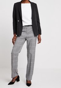 Černý dvouřadý blejzr přes bílý top, v kombinaci se šedými kostkovanými kalhotami a černými botami na nízkém podpatku. Elegantní, přizpůsobený střih.