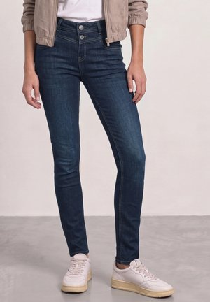 Personne portant un jean skinny bleu foncé, une veste bomber beige, des baskets blanches avec des semelles beiges, debout sur un sol gris clair avec un fond uni.