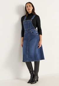 Denim hängselklänning i mörkblått, knälång, med justerbara axelband, framficka och silverfärgade knappar. Bärs med svarta knähöga stövlar.