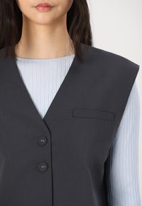 Sleeveless dark gray vest with two buttons and a small pocket, layered over a light blue textured long-sleeve top.