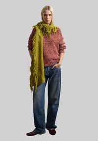 Suéter de punto en tonos rojos y multicolores, combinado con una bufanda verde texturizada. Jeans azul sueltos y zapatos burdeos completan el conjunto.