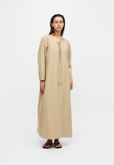 Femme portant une longue robe beige avec un détail à lacets sur le devant et des lacets assortis aux manches, debout devant un fond blanc uni.