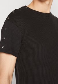 Sort ribbet t-shirt med korte ærmer, rund halsudskæring og sølvfarvede øjer på skulderen. Glat tekstur, tætsiddende design.