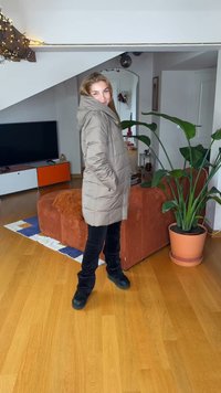 Beige polstret frakke med hætte og to sidelommer, båret over sorte bukser og sorte sko. Indendørs miljø med en plante og møbler.