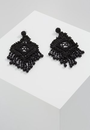 Boucles d'oreilles pendantes noires avec perles, avec un centre en forme de losange et plusieurs pampilles de perles sur une surface blanche inclinée.