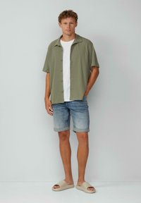 Chemise à manches courtes olive portée par-dessus un t-shirt blanc, associée à un short en jean bleu clair retroussé et des sandales glissantes beiges.