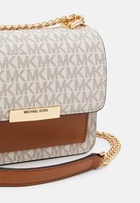 Torebka typu crossbody Michael Kors z beżowym monogramem na płótnie, brązowymi skórzanymi akcentami, złotą łańcuszkową rączką oraz plakietką z logo.