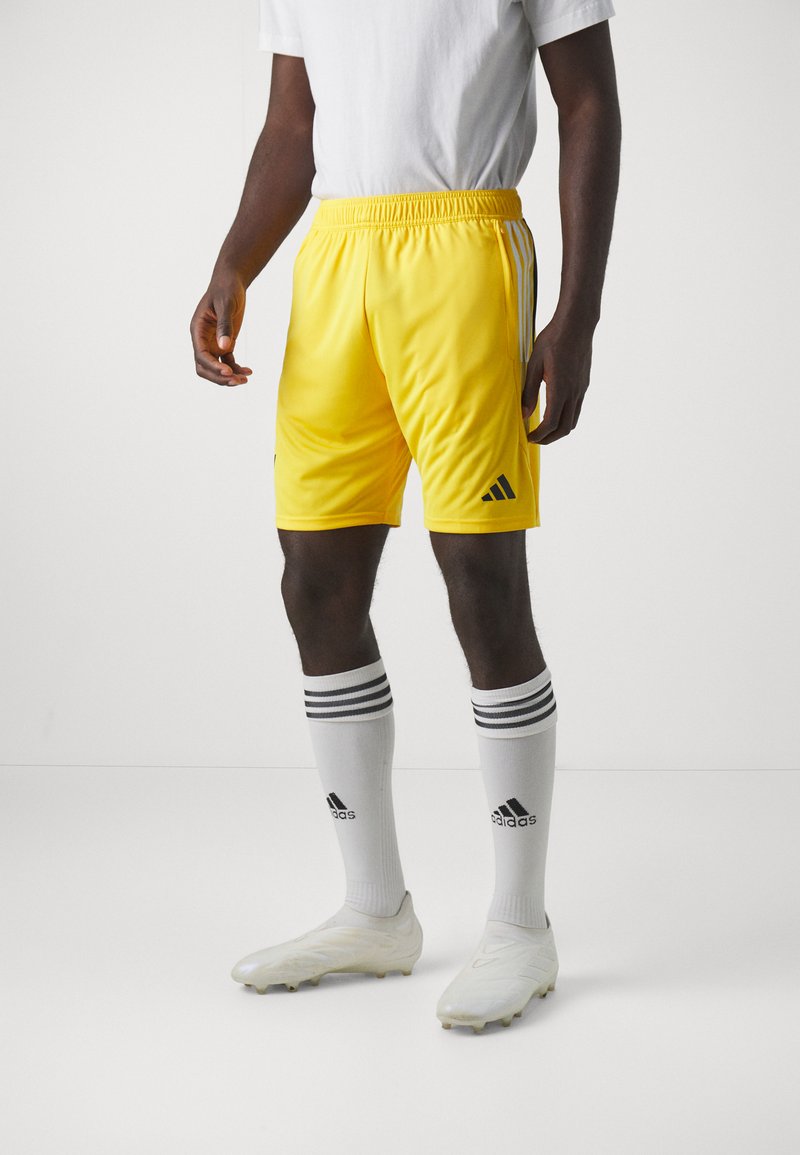 Pantalones cortos de fútbol amarillos con una cinturilla elástica, logo de Adidas en negro y tres franjas blancas en los lados; combinados con calcetas blancas hasta la rodilla y botas de fútbol.