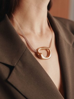 Colar com pendente dourado de forma orgânica e suave, combinado com uma corrente de malha fina, usado sobre uma peça de vestuário escura com colarinho.