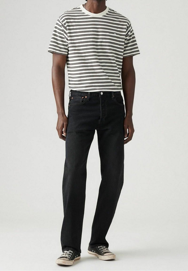 Homme portant un jean noir, un t-shirt rayé noir et blanc et des baskets basses noires, debout devant un fond uni.