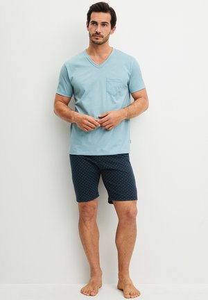 Mann steht barfuß, trägt ein hellblaues T-Shirt mit V-Ausschnitt und Tasche sowie marineblaue Shorts mit kleinen weißen Punkten vor einer weißen Wand.