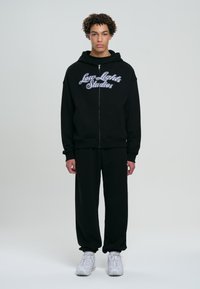 Felpa nera con zip e il testo ricamato bianco "Low Lights Studios" sul petto, abbinata a pantaloni della tuta neri e sneakers bianche.