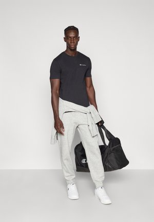 Black cotton t-shirt, gray sweatpants, white sneakers. Model holds a black duffel bag. Sweatshirt wrapped around the waist.