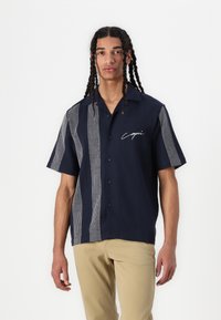Camisa de manga corta en color navy con un diseño de patrón mixto que presenta detalles a cuadros en un lado y un logotipo característico en el pecho.