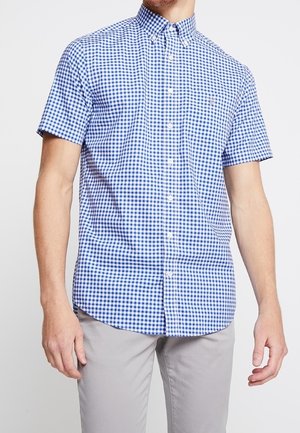Homme portant une chemise à manches courtes à carreaux bleu et blanc avec boutons et un pantalon gris clair, debout devant un fond uni.