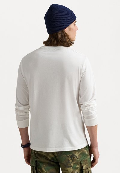 Λευκή μακρυμάνικη μπλούζα με υφασμάτινους σφιγκτήρες, συνδυασμένη με καμουφλαρισμένο παντελόνι cargo. Φοριέται με ναυτικό πλεκτό καπέλο. Πίσω όψη, λείο ύφασμα.