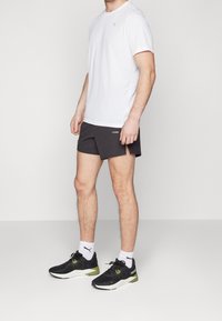 Vit t-shirt med kort ärm, mörkgrå shorts, svarta träningssneakers med gröna detaljer, och synliga vita strumpor. Lättviktsmaterial, avslappnad design.