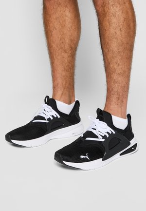 Jambes portant des baskets Puma noires et blanches avec des détails en mesh et en caoutchouc, associées à des chaussettes blanches aux chevilles, sur un sol gris clair uni.