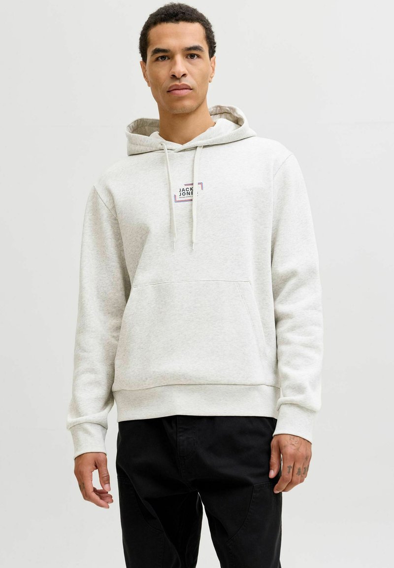Sweat à capuche gris clair en tissu doux, avec une poche avant et un patch logo sur la poitrine. Le mannequin porte un pantalon noir.