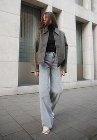 Giacca in pelle oversize verde sopra un dolcevita nero, abbinata a jeans a gamba larga grigio chiaro e sandali bianchi, in piedi su un marciapiede.