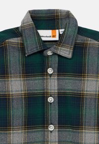 La chemise en flanelle à carreaux présente un motif à carreaux vert, gris et jaune, un devant à boutons et un col structuré avec des boutons en métal.