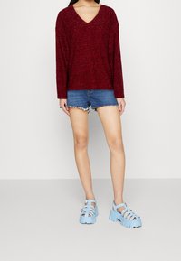 Pull bordeaux à col en V et manches longues, associé à un short en jean ; sandales compensées bleues avec un design à brides et des semelles texturées.