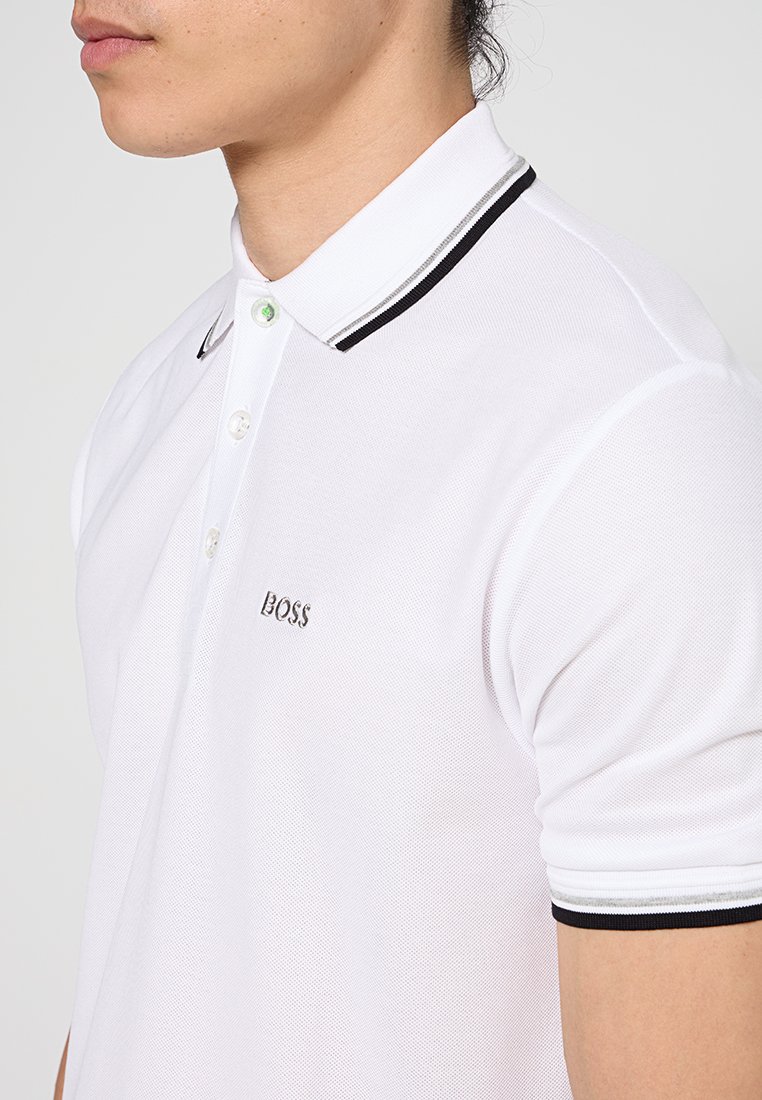 Camisa polo blanca hecha de tejido texturizado con un cuello de rayas en blanco y negro, tres botones y un pequeño logo bordado.