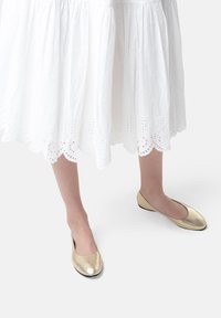 Zapatos planos dorados con un acabado metálico suave, punta redondeada y tacón bajo, combinados con una falda blanca de puntilla con dobladillo en forma de concha.