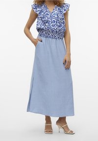 Ljusblå maxiklänning med mönstrad, volangprydd ärmlös topp, resår i midjan, slits framtill och sidofickor; bärs med beigea klackskor.