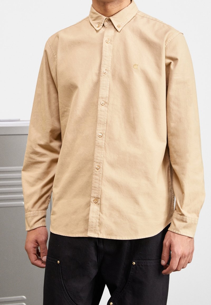 Camisa de botón de manga larga en algodón beige, con cuello clásico y logotipo bordado en dorado en el pecho. Textura suave, ajuste estándar.