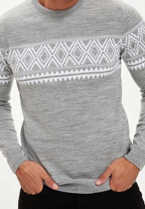 Uomo che indossa un maglione grigio chiaro con motivo geometrico bianco a diamanti e triangoli sul petto e sulle braccia, mani nelle tasche.
