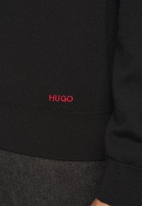 Pull en maille noire avec une texture côtelée et un logo rouge "HUGO" à l'ourlet. Le pull est ajusté avec des manches longues.