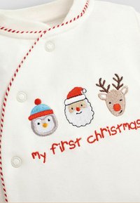 Un body de algodón blanco con ribete rojo; presenta un pingüino bordado, Papá Noel y reno; el texto dice "mi primera Navidad" en rojo.