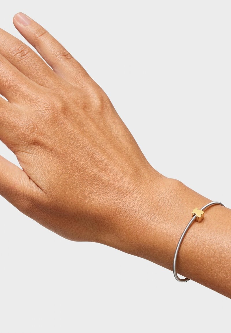Bracciale in metallo argento con accento di croce dorata, caratterizzato da una superficie liscia e un design minimalista, indossato su una mano.