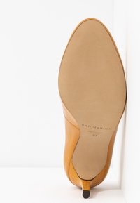 Żółty skórzany but na wysokim obcasie z ostro zakończonym noskiem i smukłym obcasem. Podeszwa jest gładka, widnieją na niej informacje o marce i rozmiarze.
