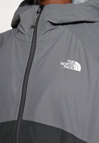 Jachetă gri tip windbreaker cu fermoar pe jumătate în față, glugă și un panou inferior de culoare neagră în contrast. Logo notabil pe piept în alb.