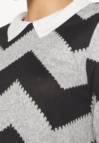 Personne portant un pull tricoté à zigzag gris et noir avec un col pointu blanc, montrant le cou et la partie supérieure de la poitrine.