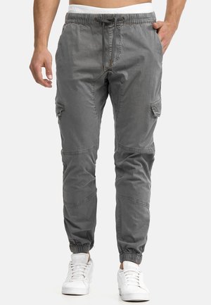 Homme portant un pantalon cargo jogger gris avec des poignets élastiques et une taille à cordon, associé à des baskets blanches sur un fond uni.